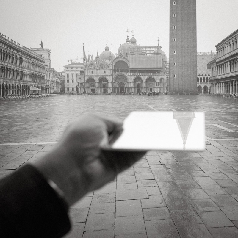 A hand holds a small mirror over Piazza San Marco, reflecting the campanile above the rain-wet square.