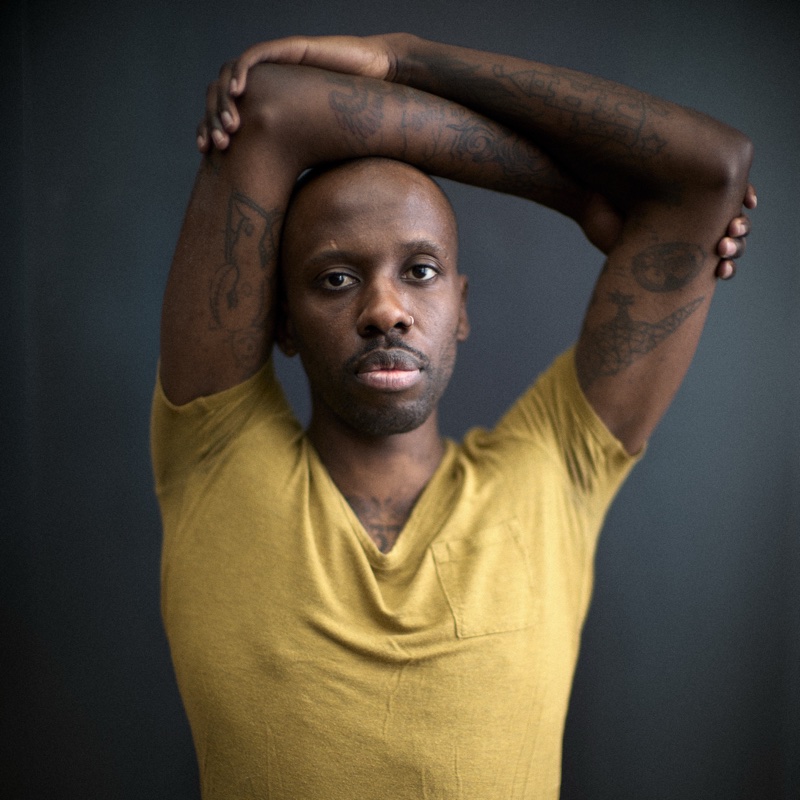 Bald man with tattooed forearms folded atop his head, in a mustard V-neck tee against a gray backdrop.