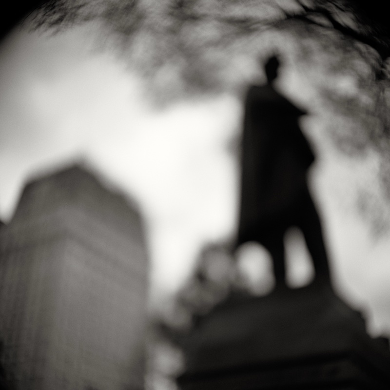Out-of-focus statue of a standing figure rises above a gravestone, framed by bare branches.