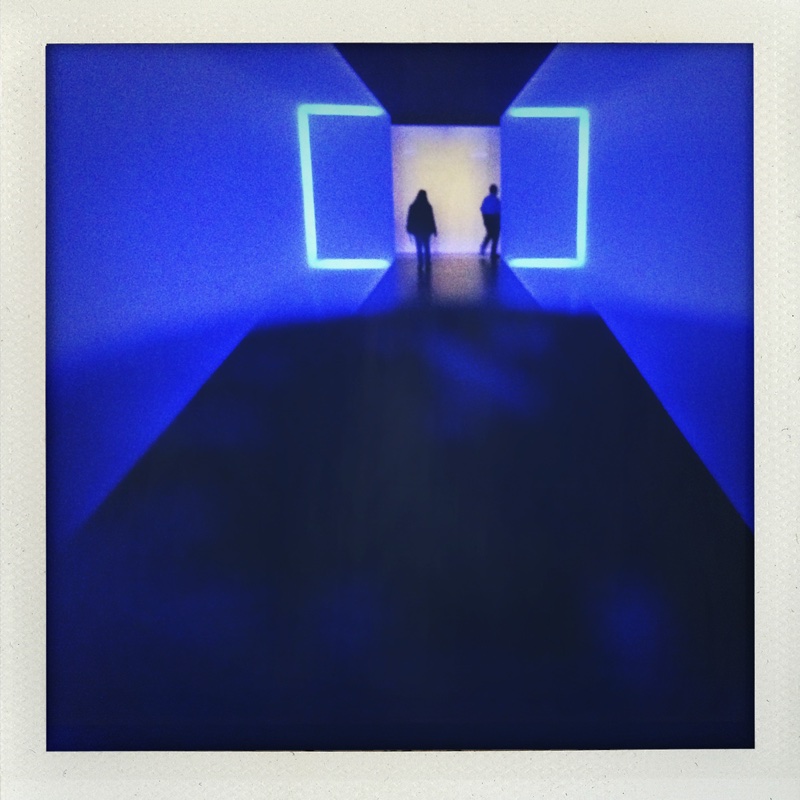 Two visitors silhouetted in a deep-blue corridor installation, walking toward a glowing doorway framed by neon outlines.