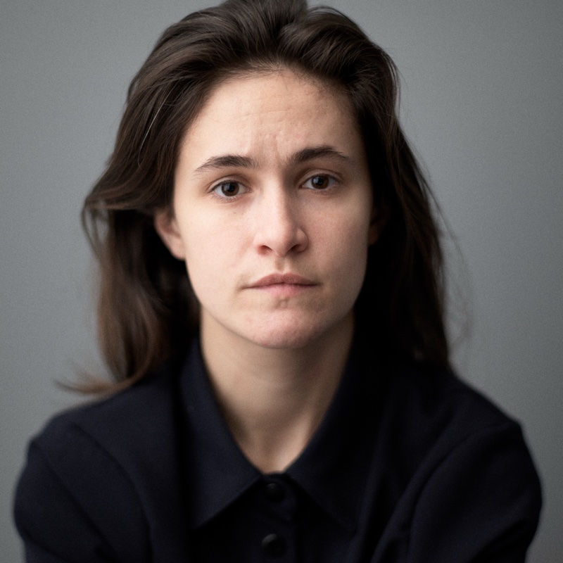 A young woman with long dark hair, in a dark collared shirt, faces the camera against a gray wall.